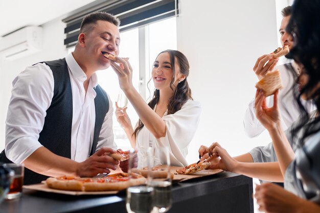 Crop portrait of pretty bride woman feeds the groom with pizza in the kitchen with guests friends of brides A loving couple celebrating engagement waiting for wedding ceremony
