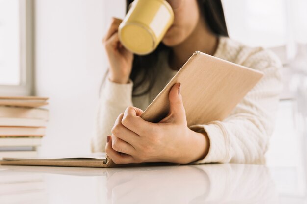 Crop woman drinking and reading at table