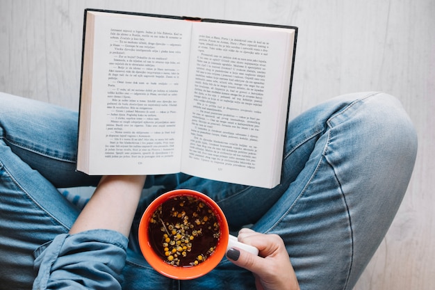 Crop woman drinking tea and reading book