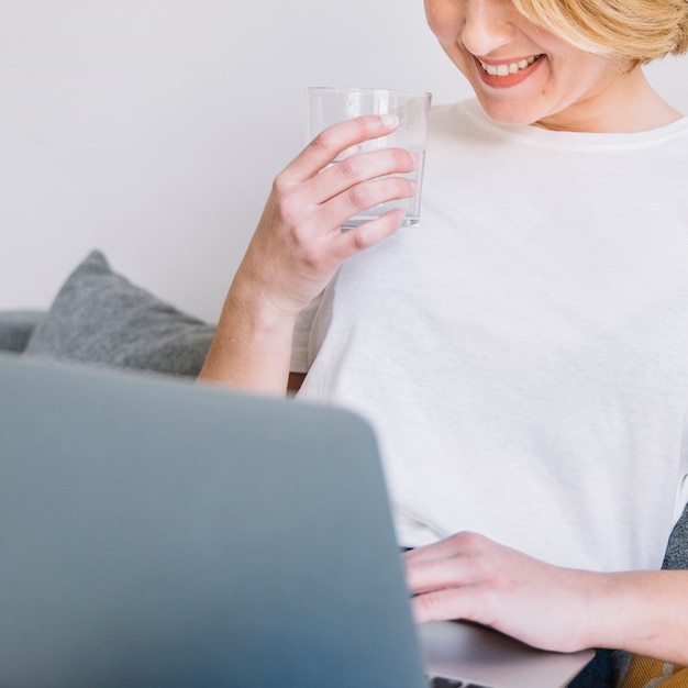 Crop woman drinking water and using laptop