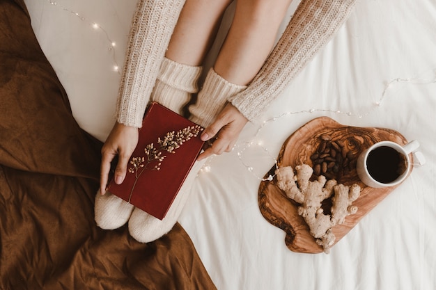 Crop woman putting book on legs near tea and snacks