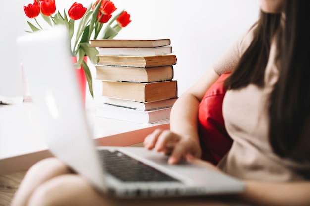 Crop woman using laptop near flowers and books