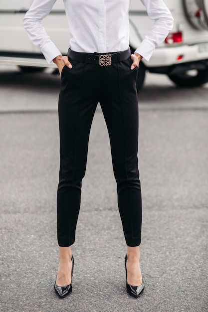 Croppe photo of fashionable and stylish lady dressed in black pants and white blouse posing on the background of a car outdoors