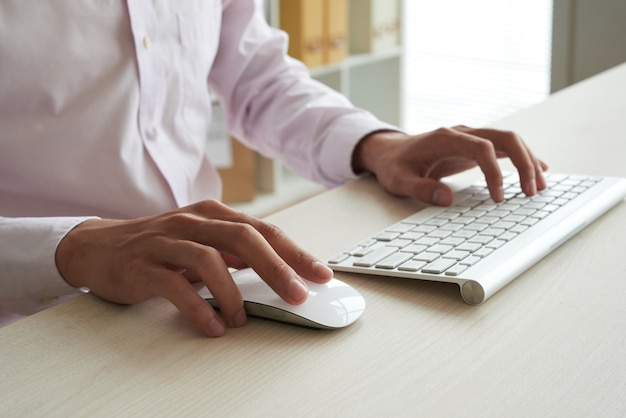Обрезанный анонимный человек, вычисляющий на белой клавиатуре и использующий белую мышь