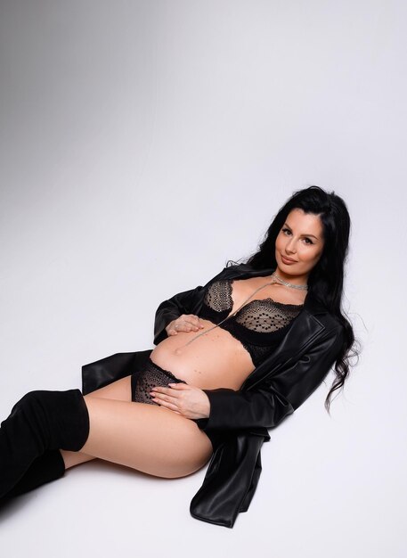 Cropped of attractive pregnant woman with professional makeup and pendant on neck wearing in black leather jacket and laced underwear touching belly while looking at camera and lying in studio