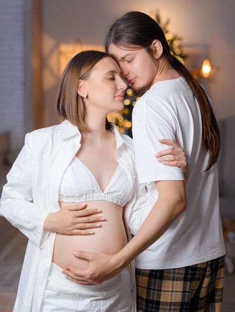 妊娠中の女性の頭に寄りかかって、背景にクリスマスの装飾が施された子供部屋に立っている間、腹に触れるパジャマを愛する男に目を閉じて美しい若いカップルのトリミング