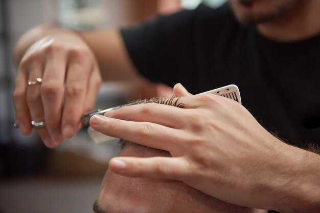 Обрезанный снимок профессионального парикмахера, который использует ножницы и расческу во время стрижки своего клиента.
