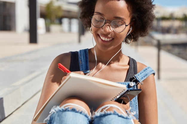 L'immagine ritagliata di attraente e allegra giovane artista femminile prende appunti creativi nel blocco note, indossa gli occhiali, gode di una bella traccia negli auricolari collegati al telefono cellulare, ha un'espressione positiva.