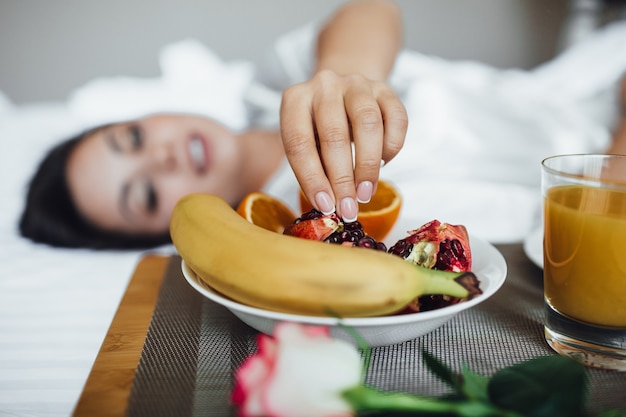 Cropped image of the beautiful brunette girl in the morning, next to croissant, orange juice and banana pomegranate on the tray and rose