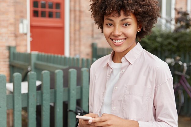 Cropped image of black woman with Afro haircut, uses mobile phone, satisfied, poses outdoor in private sector near her house,