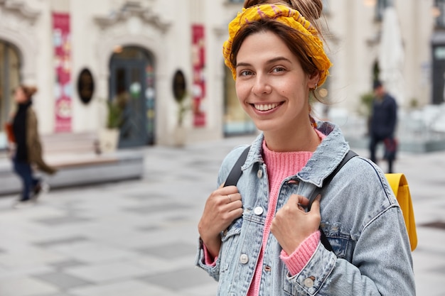 Cropped image of cheerful young Caucasian woman strolls across city with small rucksack, wears yellow headband and denim jacket