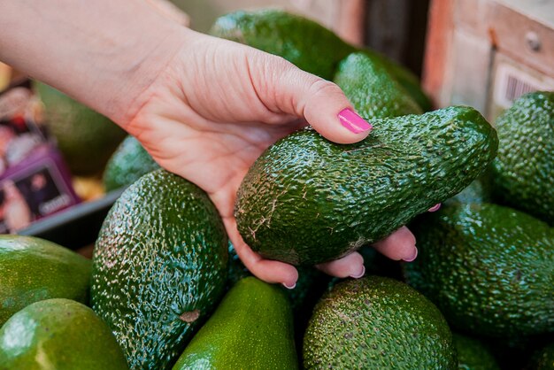 Обрезанное изображение клиента, выбирающего авокадо в супермаркете. Закрыть женщина рука, проведение авокадо на рынке. Концепция продажи, покупки, продуктов питания, потребительства и людей