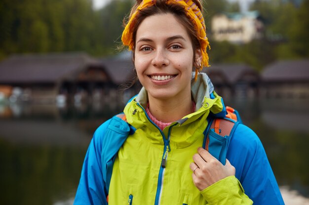 Cropped image of delighted woman with toothy smile, wears casual jacket, carries rucksack