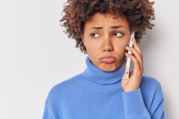 Обрезанное изображение недовольной кудрявой женщины кладет губы в сумку, чувствует себя грустно, смотрит несчастно в сторону, имеет покрытие телефона, держит сотовый телефон рядом с ухом, одетая в синюю водолазку, изолированную на белой стене