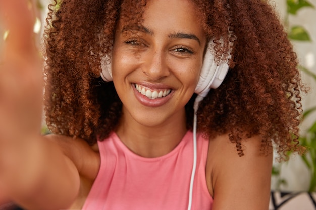 Cropped image of glad mixed race black woman with crisp hair, makes selfie portrait, listens audio track in headphones, being in high spirit, has spare time afer classes, enjoys exciting audio book