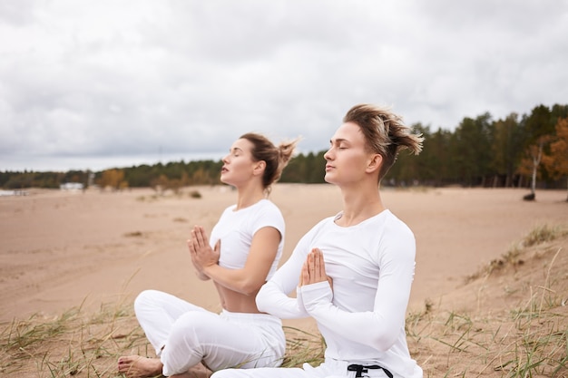 Free photo cropped image of handsome guy practicing meditation with blonde woman, sitting on sand in lotus pose, closing eyes, having peaceful facial expressions.