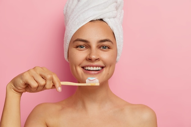 Cropped image of happy European woman brushes teeth, holds toothbrush with toothpaste, wears wrapped towel on head, has healthy fresh skin, stands naked
