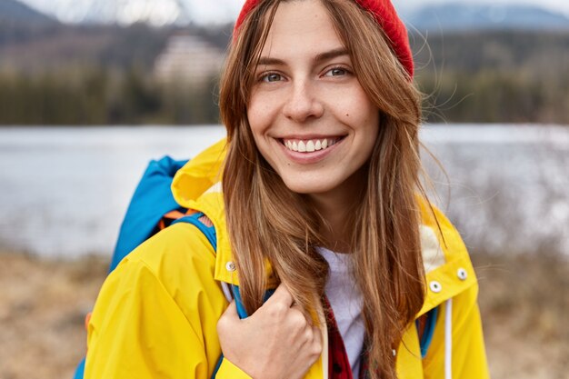 Cropped image of lovely cheerful European woman has broad tender smile, long straight hair, wears red hat