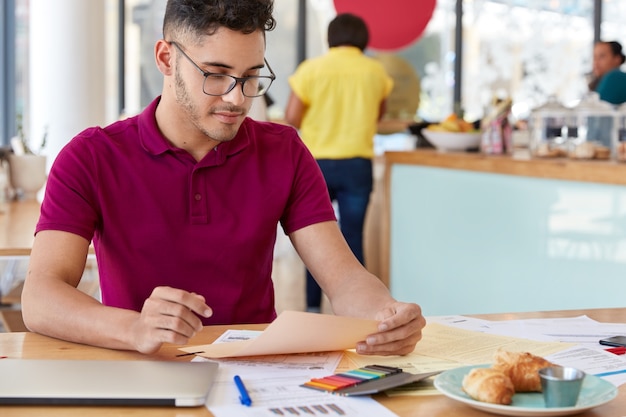 Cropped image of male freelancer studies financial report, makes project, dressed in casual clothes, sits at desktop with colourful stickers, croissants, wears casual outfit. Paperwork concept