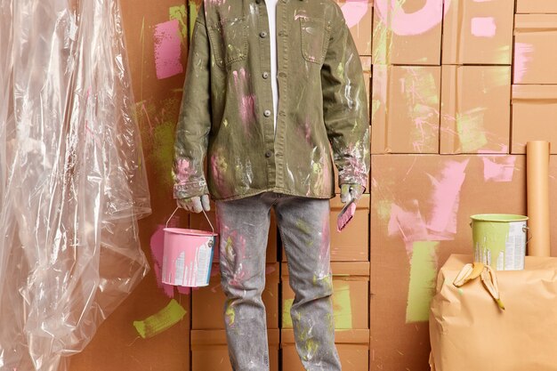 남자 화가의 자른 이미지는 분홍색 페인트 양동이를 보유하고 브러시는 캐주얼 셔츠와 청바지를 입고 방에 벽을 칠하는 집 마감의 빠른 수리를 수행합니다. 유지 보수 및 주택 개선 개념
