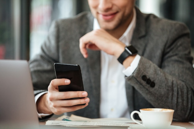 Обрезанное изображение счастливого бизнесмена, сидя за столом в кафе и с помощью смартфона