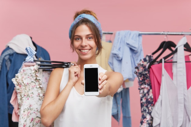 Cropped image of positive young female dressed casually, doing shopping while standing with hagers of garment, holding modern smart phone in hand with blank screen