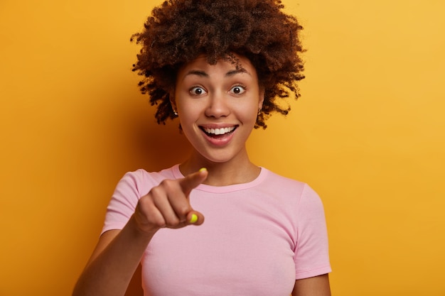 Cropped image of pretty joyful woman with toothy smile points directly , sees something amazing in front, wears t shirt, has curious happy expression, isolated on yellow wall.