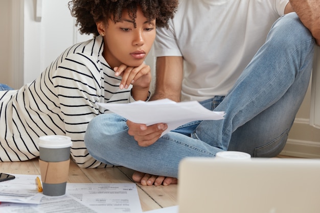 Free photo cropped image of serious black woman and her partner look attentively at documents, develop startup project, work at home, drink hot beverage, work with laptop computer, going to present business plan