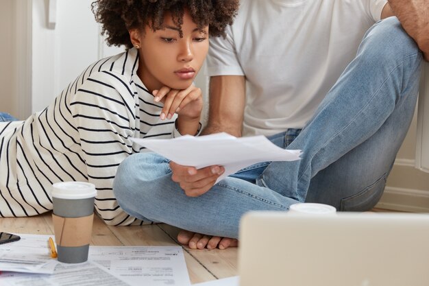 Cropped image of serious black woman and her partner look attentively at documents, develop startup project, work at home, drink hot beverage, work with laptop computer, going to present business plan