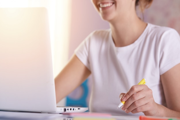 Free photo cropped image of smiling positive woman being busy, using white laptop, surfing net, working online, being freelancer
