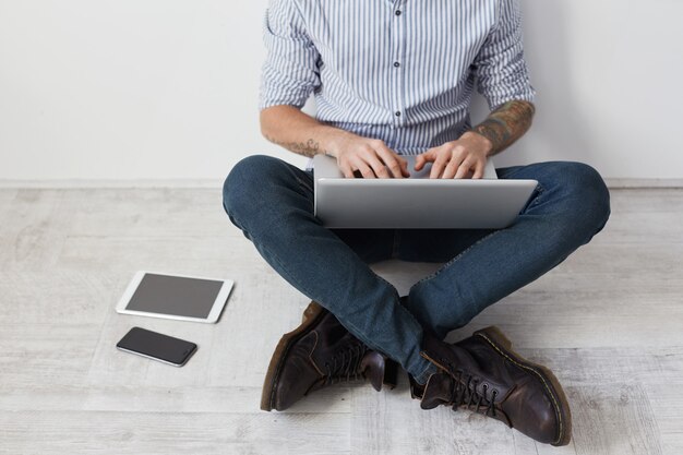 Cropped image of stylish tattooed hipster guy wears shirt, jeans and boots, sits crossed legs on floor,