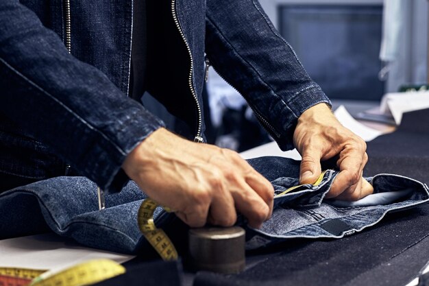 Cropped image of tailor makes measurements of jeans with a meter at sewing workshop.