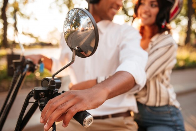 Cropped image of young african couple rides on modern motorbike in park and looking to each other. Blur image