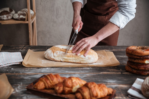 Cropped image of young man baker cut the bread.