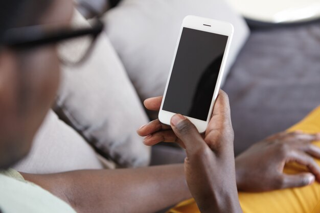 Cropped indoor image of unrecognizable dark-skinned man relaxing on couch in living room, using home wi-fi on modern mobile phone