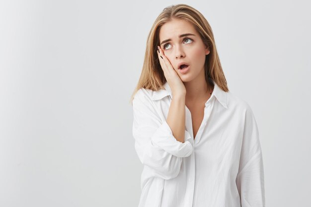 Cropped isolated shot of tired young blonde female touching her face and wearing white shirt, mouth wide open, looking annoyed and bored