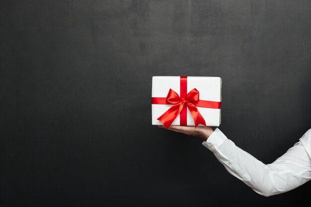 Cropped male hand holding white present box with red bow, isolated over dark gray wall