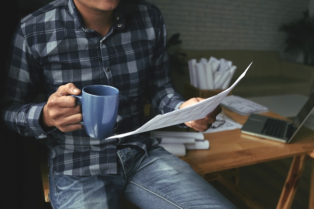 Free photo cropped man checking blueprint while having tea