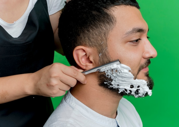 Cropped pfoto of young professional hairdresser woman in apron putting shaving foam and shaving beard of man client over green background