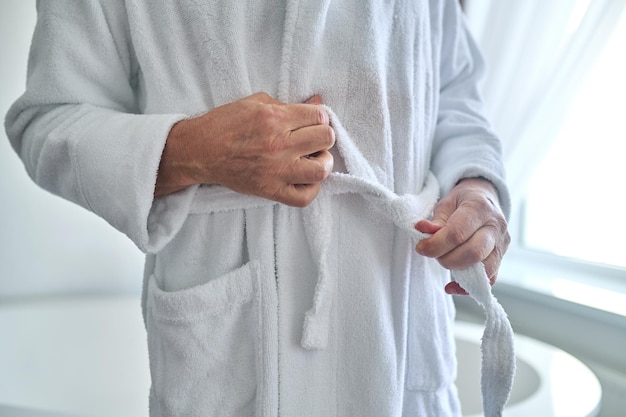 Cropped photo of a male standing near the bathtub while tying the belt of his towelling robe