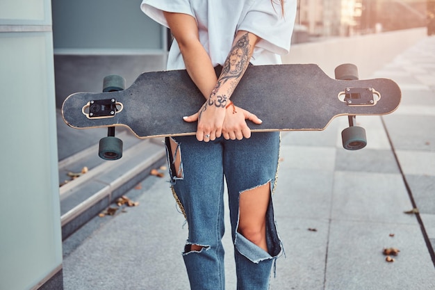 無料写真 白いシャツと破れたジーンズに身を包んだ若いヒップスターの女の子のトリミングされた写真は、超高層ビルの近くでポーズをとっている間スケートボードを保持しています。