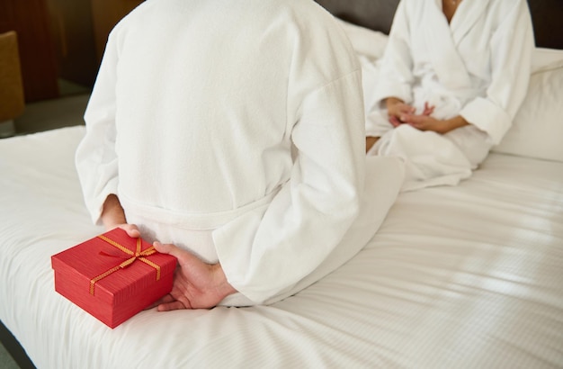 ベッドの上で恋人の前に座って赤いギフトボックスを隠している白いテリー織りのバスローブを着た認識できない男性のトリミングされた写真は、彼女をバレンタインデーの驚きにしています。恋人たちは休日に贈り物をします Premium写真