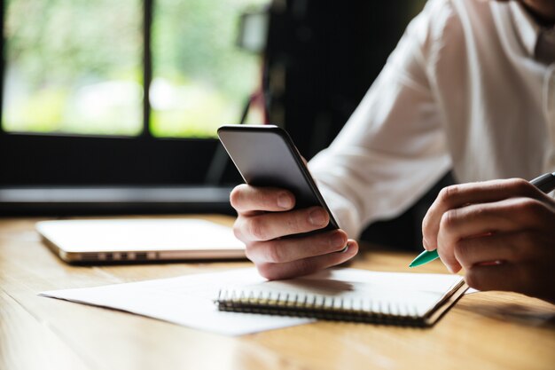 Cropped photo of young man in white shirt holding smartphone, while resting after paperwork