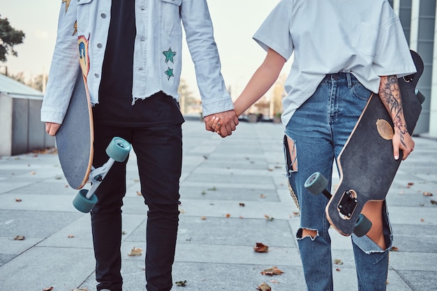 Cropped photo of a young trendy dressed skaters hold hands while standing near skyscraper.