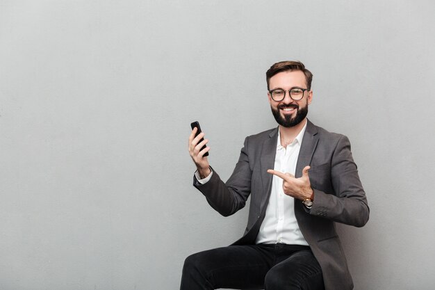 Cropped picture of happy man in eyeglasses looking on camera while sitting on chair and pointing on his mobile phone, isolated over gray