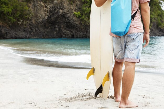 Cropped portrait of barefooted young surfer standing on sandy beach against high rocky shore with vegetation, carrying his white surfboard, ready to hit waves