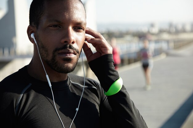 Cropped portrait of black sportsman sitting on pavement in deep thoughts, listening to motivative audiobook in his headphones, touching his head, looking confident and concentrated during workout