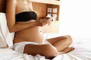 Free photo cropped portrait of tanned young expectant mother dressed in underwear holding smart phone, using high-speed internet connection at home or in hotel room