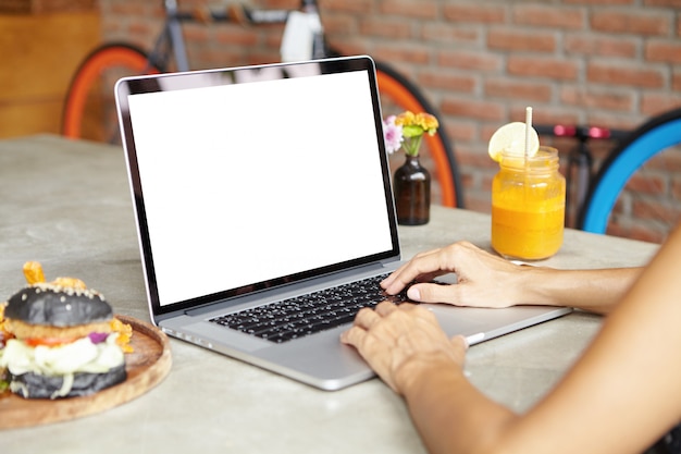 Cropped rear portrait of self-employed woman using laptop computer for distant work while having meal at cafe with brick wall. Female designer working on her project on notebook pc during lunch