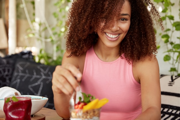 분홍색 캐주얼 티셔츠를 입은 곱슬 검은 머리를 가진 사랑스러운 여성 모델의 자른 샷은 디저트를 먹고 광범위하게 미소 짓습니다. 혼합 된 경주 젊은 아프리카 계 미국인 여자 카페 인테리어에 대 한 포즈.